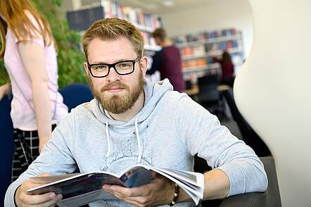 Foto: Ein Studierender sitzt in der Bibliothek und liest eine Zeitschrift. 