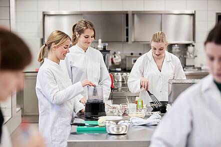 Foto: Studentinnen in weißen Kitteln stehen an einer Kochstation in einem Labor und arbeiten mit Lebensmitteln