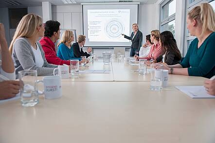 Foto: Studierende sitzen an einem langen Tisch und nehmen an einem Präsenzseminar teil.