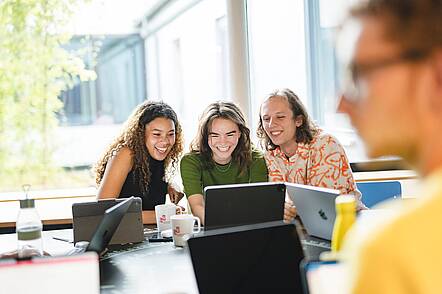 Foto: Zwei Studentinnen und ein Student sitzen an einem Tisch und blicken gemeinsam in einen Laptop