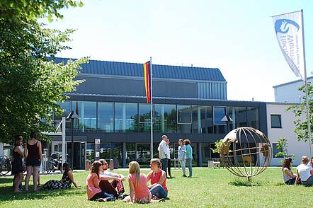 Foto: Studierende sitzen im Sommer vor dem Hochschulgebäude