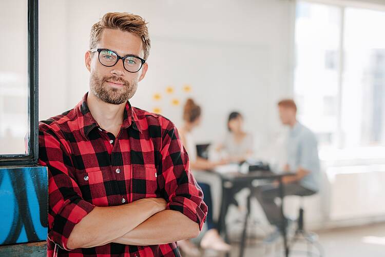 Foto: Studierender, der mit verschränkten Armen dasteht, während sich im Hintergrund weitere Studierende unterhalten.