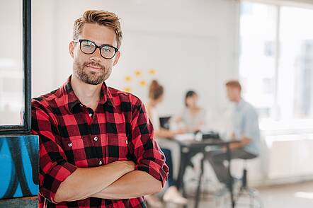 Foto: Studierender, der mit verschränkten Armen dasteht, während sich im Hintergrund weitere Studierende unterhalten.